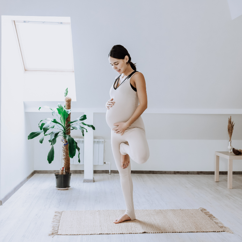 A pregnant woman standing in a yoga pose on a yoga mat in a room with a window and a plant