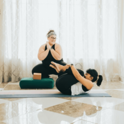 A plus-sized female yoga instructor instructing another woman with her knees to her chest on a yoga mat with a bolster
