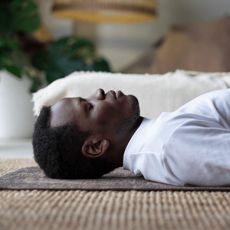 A Black man lying on a yoga mat on the floor with his eyes closed.