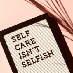 An image of a white letter board with a black frame that reads "self care isn't selfish" in black letters, set against a pink background with a shadow of a palm branch