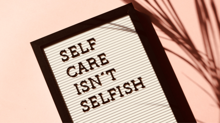 An image of a white letter board with a black frame that reads "self care isn't selfish" in black letters, set against a pink background with a shadow of a palm branch